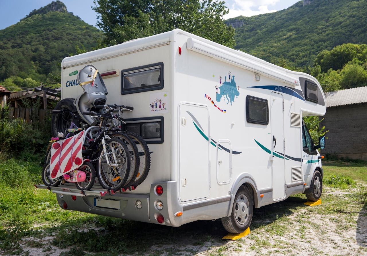 our-stories An RV with bicycles attached in a scenic outdoor setting, surrounded by mountains and greenery.