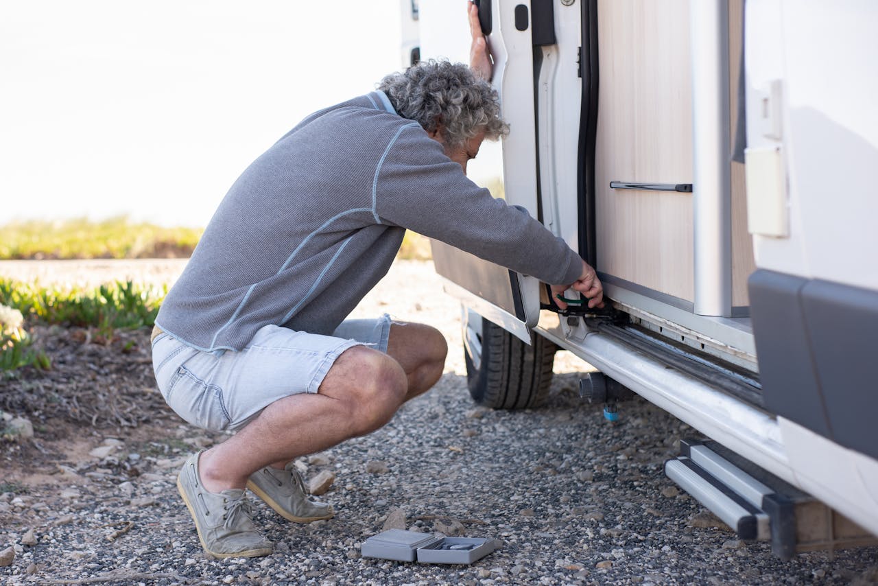 Senior man fixing camper van door mechanism outdoors. Emphasizes travel and hands-on problem-solving.